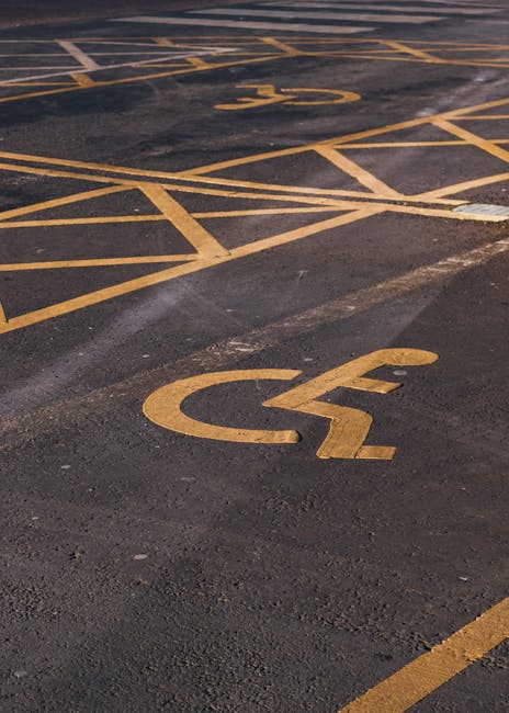 Close-up of a yellow-painted wheelchair accessible parking symbol on an asphalt surface, located in a designated parking area. The symbol depicts a stylised image of a person sitting in a wheelchair,
