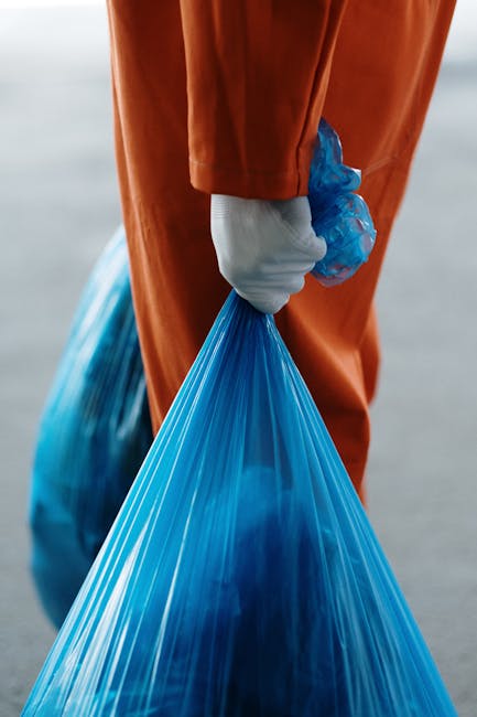 A sanitation worker wearing a high-visibility yellow vest and black trousers is positioned at the rear of a large red waste collection vehicle, engaged in the loading or unloading process of trash. Th