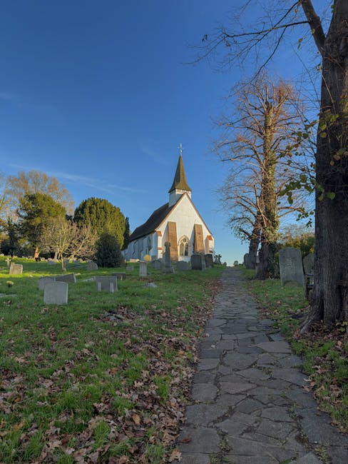 A narrow, uneven cobblestone path leading uphill through a grassy area with fallen leaves, situated in a churchyard. On the right side of the path, there are several leafless and partly leafy trees, some with green foliage further back. To the left, there are multiple small gravestones and a few larger monuments scattered across the lawn. A white, small church with a pointed steeple and a shingled roof stands at the top of the incline under a clear blue sky, with sunlight illuminating its facade. The scene is peaceful and well-maintained, reflecting a typical historic churchyard setting, with a focus on the home relocation context related to house removals and moving logistics. Man and Van Harlington is involved in the transportation and packing processes implied by the setting, with the area serving as a potential loading zone for furniture and belongings during a house move.