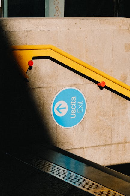 A close-up view of a concrete staircase with a yellow safety handrail and black rubber strip along the edge, located outdoors near an entrance area. The staircase features a circular blue sign with the word 'Uscita' and an upward-pointing arrow, indicating an exit, attached to the side wall. The scene is lit by natural daylight, with some shadows cast on the wall. Visible around the staircase is a metallic drainage grate at the base, suggesting a surrounding environment where furniture and household items are likely being moved during a home relocation or furniture transport process. The image is relevant to relocation services provided by Man and Van Harlington, particularly in the context of loading or accessing exits during house removals near Hayes and Harlington station.