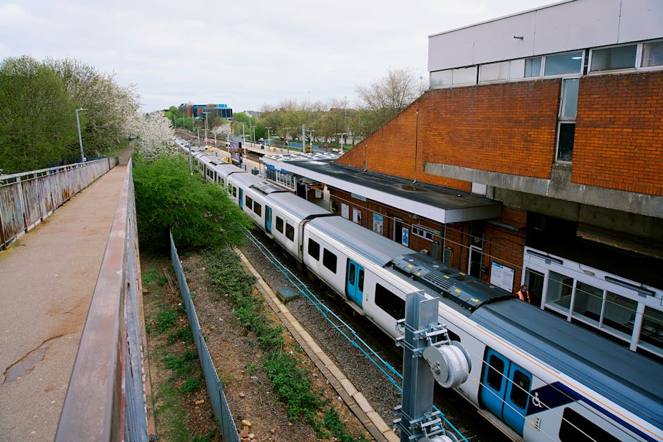 A view from a high-level pedestrian walkway overlooking a train station platform with a modern, multi-carriage train parked alongside it. The train is predominantly silver with blue accents around the doors and windows. The platform has a small shelter structure with a flat roof and a notice board attached, positioned adjacent to the station building constructed of red brick and concrete. On the platform, there are no visible passengers, but the scene suggests an ongoing home relocation or furniture transport process supported by Man and Van Harlington. To the left, a metal railing runs parallel to the walkway, which is paved with a mixture of asphalt and concrete, bordered by some greenery and trees. The environment appears to be during daylight with overcast skies, and the scene captures part of the loading or unloading activity involving moving boxes and furniture, possibly being prepared for transportation as part of the house removals service.
