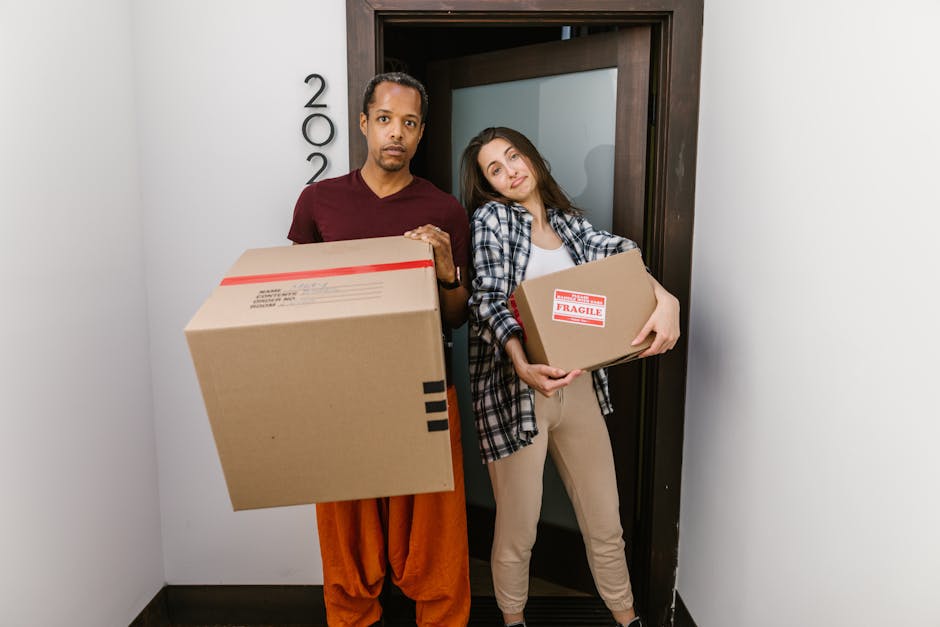 A man and a woman are standing inside a residential doorway, each holding a cardboard box as part of a home relocation process. The man, dressed in a maroon t-shirt and orange trousers, is holding a large cardboard box, while the woman, wearing a plaid shirt over a white top and beige trousers, is carrying a smaller box labeled with a red 'Fragile' sticker. They appear to be preparing for a move, possibly with the assistance of Man and Van Harlington, as they stand close to the doorway with the door open behind them. The scene is well-lit, with plain white walls on either side and a dark wooden door frame, suggesting it is daytime and part of a local house move in the Heathrow area. The boxes are made of standard brown cardboard, and the woman’s box shows some packing tape along the edges, indicating secure packing for transport. This image captures the typical loading phase of a small local move, where furniture and belongings are prepared for transport, reflecting professional packing and handling practices aligned with local house removal services.