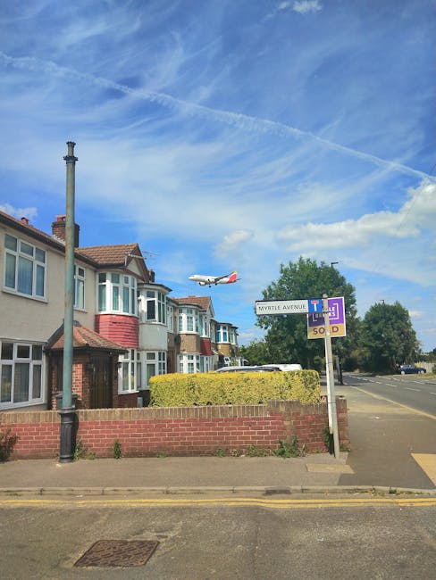 A clear daytime view of a residential street in the Heathrow area, showing a row of typical UK semi-detached houses with bay windows, brickwork, and small front gardens. An airplane is captured mid-flight against a bright blue sky with wispy contrails. The scene includes a street sign indicating Myrtle Avenue and nearby parking regulations, along with a lamp post and a small hedge bordering the sidewalk. The street appears calm with no visible moving vehicles or pedestrians. This setting offers context for local house removals or home relocation services by illustrating an ordinary residential environment, relevant to moving logistics in the Harlington area, as provided by Man and Van Harlington.