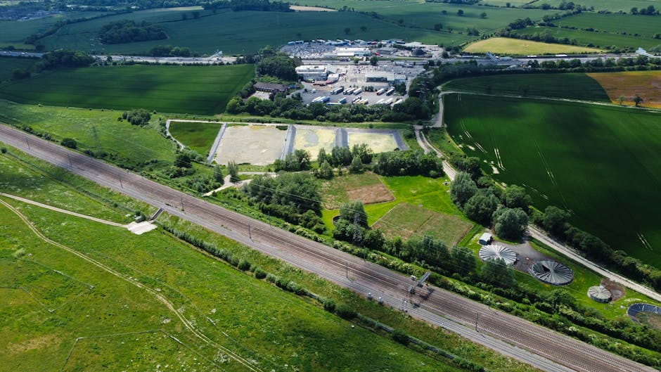 An aerial view of a property with a green garden and surrounding fields, with various moving boxes and furniture covered in plastic and blankets visible near the house's entrance. A large van or lorry is parked close by, with a few workers actively lifting and loading cardboard boxes and furniture onto the vehicle within the property’s driveway. The scene includes equipment such as trolleys and straps used for safe transport, and the open doorway reveals part of the interior preparing for home relocation. Outside, the nearby railway tracks run parallel to the property, with lush green grass and hedges lining the paths. In the background, there is an industrial or commercial area with buildings, parking lots, and additional open spaces, all under bright daylight. This image supports concepts of packing and moving logistics, furniture transport, and the process of house removals, as detailed by Man and Van Harlington for Hillingdon Council waste rules compliance.