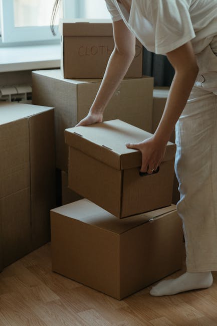 Interior view of a room during a home relocation, featuring several cardboard boxes used for packing and moving. One open box in the foreground contains crumpled grey packing fabric or clothing, likely used to protect belongings during transport. Behind it, there are three more closed cardboard boxes, two of which are marked with red and white labels, indicating they are prepared for removal. The boxes vary in size and are placed on a concrete or similar hard flooring surface, with some boxes resting near a doorway and others on the main floor. This scene captures the packing process integral to furniture transport and house removals, with visible materials such as cardboard, packing fabric, and the temporary storage environment. The lighting appears natural, typical of a residential interior, and the setting exemplifies the organization involved in professional removals by [COMPANY_NAME], preparing belongings for a smooth and efficient move related to house or flat relocation services.