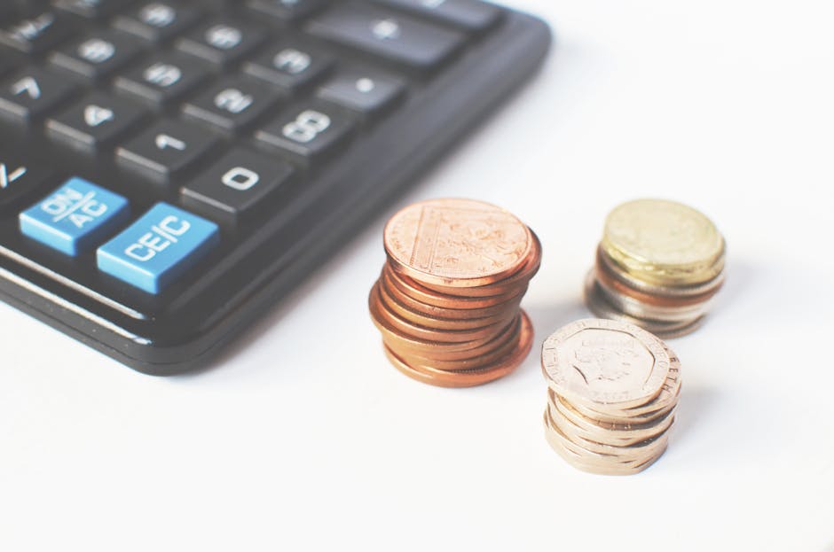 A close-up image showing a black calculator with blue and white buttons positioned on the left side of the frame, partially visible. To the right of the calculator, there are several stacks of coins, 