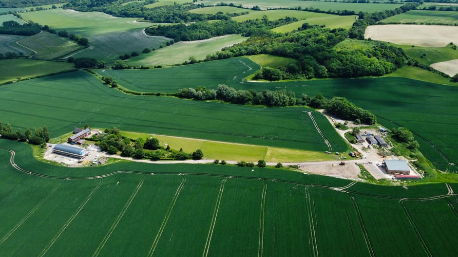 An aerial view of a rural landscape featuring rolling green fields with distinct crop lines, interspersed with patches of trees and small wooded areas. In the foreground, there are several large industrial or agricultural buildings surrounded by paved and gravel areas, possibly farm storage or workshops, with a few vehicles parked nearby. A narrow driveway or access road connects these buildings, extending towards the background through the countryside. The landscape shows a mix of open farmland and clusters of trees, under a clear sky with good natural lighting. This scene illustrates the expansive rural environment typical for house removals and relocation services, highlighting the importance of careful logistics in rural property moves.