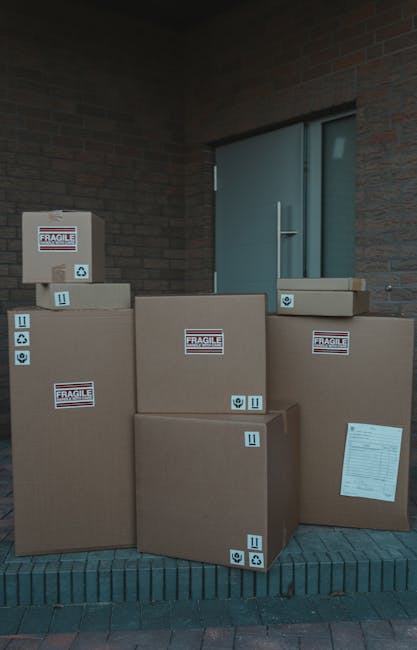 A collection of six large cardboard boxes of varying sizes, all labeled with 'Fragile' stickers, are stacked outside a building near a doorway. The boxes are securely sealed with packing tape and some feature small handling and orientation symbols, indicating careful handling. The packages are arranged on a paved area at the foot of a brick wall, adjacent to a modern door with a metal handle, and appear to be prepared for a home relocation or furniture transport process. Behind the boxes, part of a window or glass panel is visible, set into the building's exterior. The scene suggests a professional removals service, such as Man and Van Harlington, is engaged in packing or loading items for a house move, with the boxes ready for transportation to a new residence, supporting efficient moving logistics, packing, or furniture transport tasks related to same-day removals in Harlington.