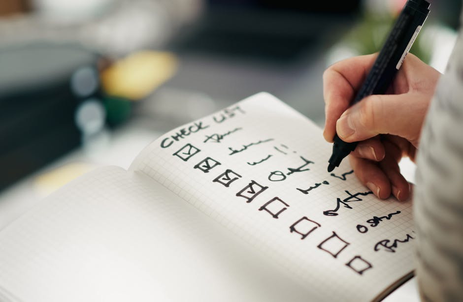 Close-up image of a person’s hand holding a black marker and writing a house removal checklist in a grid notebook. The checklist includes items such as 'check list,' 'arrival,' 'pack,' and 'loading.' The person is inside a property, with a glimpse of a black storage box and some yellow objects, possibly furniture or household items, visible in the blurred background. The scene captures the preparation and organisation process involved in home relocation, with a focus on packing, loading, and logistical steps. The lighting is natural, highlighting the careful documentation necessary for a smooth moving and furniture transport process. Man and Van Harlington, a company specialising in removals, often assist clients with packing and moving logistics, as reflected by the detailed planning shown in this image.